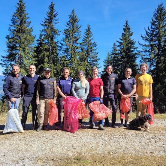 Eine Gruppe von Menschen steht auf einem Schotterweg, hält Plastiktüten in den Händen und hat Bäume und einen Berg im Hintergrund. Sie sind in Outdoor-Kleidung gekleidet, einige mit Mützen.