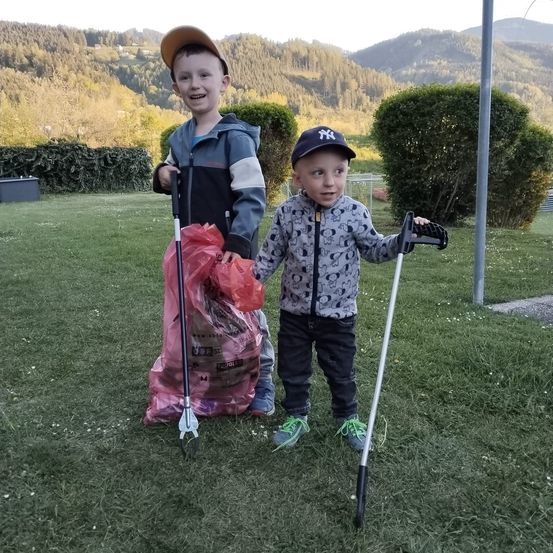 Zwei kleine Jungen, einer mit einer Harke und der andere mit einem Stock, stehen auf einem Rasen mit einem rosa Beutel voller gesammelter Gegenstände. Dahinter ist ein Busch und Berge zu sehen.