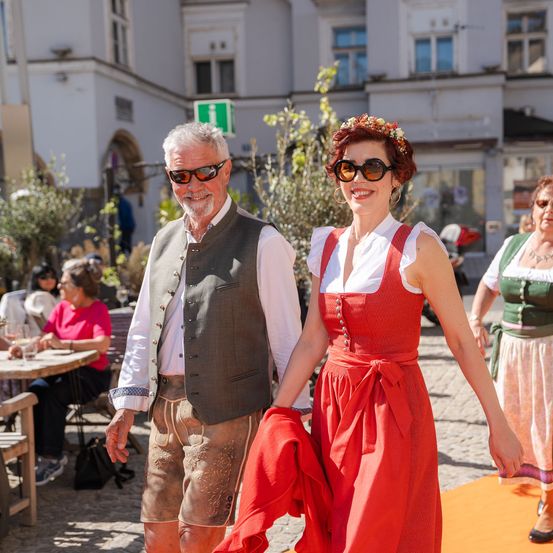 Ein älterer Mann und eine Frau in traditioneller Kleidung gehen lächelnd zusammen. Sie befinden sich auf einem orangefarbenen Teppich, mit Menschen, die am Hintergrund an Tischen sitzen.