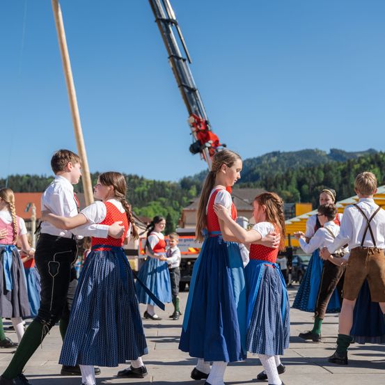 Mehrere Kinder in traditionellen Kostümen tanzen auf einem Platz, mit Bergen und einem Kran im Hintergrund.