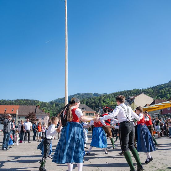 Eine Gruppe von Menschen tanzt in traditioneller bayerischer Kleidung um einen Fahnenmast, während Zuschauer von den Seiten zusehen. Im Hintergrund befinden sich Gebäude und Berge.