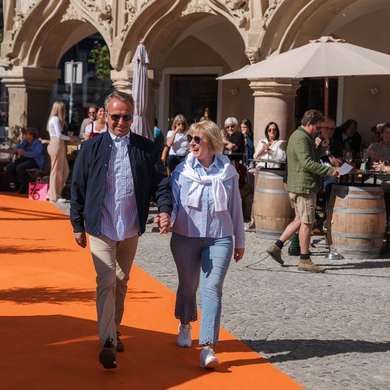 Ein Mann und eine Frau gehen auf einem orangefarbenen Teppich in einem Außenbereich. Mehrere Personen sitzen an Tischen, und einige stehen. Der Bereich hat einen gewölbten Eingang, und es gibt Holzfässer.