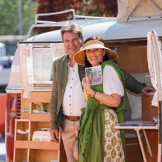 Ein Mann und eine Frau stehen vor einem alten VW-Bulli, lächelnd. Der Mann hält ein Buch, die Frau eine Karte. Der Bulli ist geöffnet und zeigt Regale und einen Tisch.