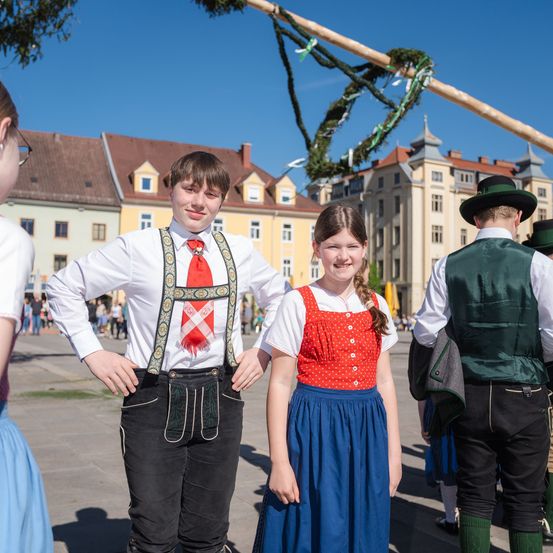 Eine Gruppe von Menschen in traditioneller Kleidung, möglicherweise bei einem Fest, steht draußen mit Gebäuden im Hintergrund. Ein Junge und ein Mädchen lächeln.