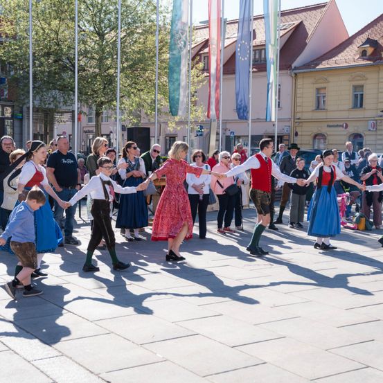 Eine Gruppe von Menschen in traditioneller bayerischer Kleidung tanzt auf einem Stadtplatz, umgeben von Zuschauern und Gebäuden mit Fahnen.