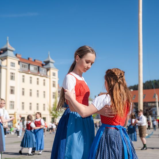 Zwei Mädchen in traditioneller deutscher Kleidung tanzen auf einem Stadtplatz, umgeben von anderen Tänzern und einem historischen Gebäude unter klarem Himmel.