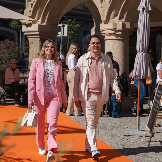 Ein Mann und eine Frau in passenden rosa Outfits gehen auf einem orangefarbenen Teppich in einer historischen Umgebung.
