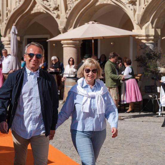 Ein Paar, beide mit Sonnenbrillen, geht Hand in Hand auf einer Kopfsteinpflasterstraße vor einem prächtigen Gebäude. Im Hintergrund sind Menschen versammelt, einige sitzen, andere stehen, eine Person hält ein Getränk.