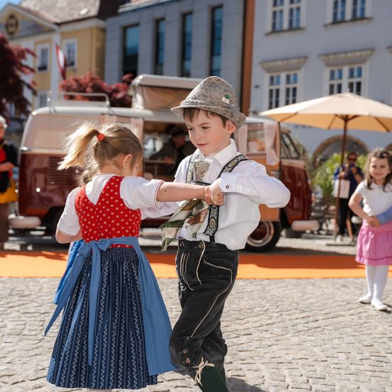 Zwei Kinder tanzen in traditioneller deutscher Kleidung auf einem Stadtplatz, mit Gebäuden und einem Van im Hintergrund.