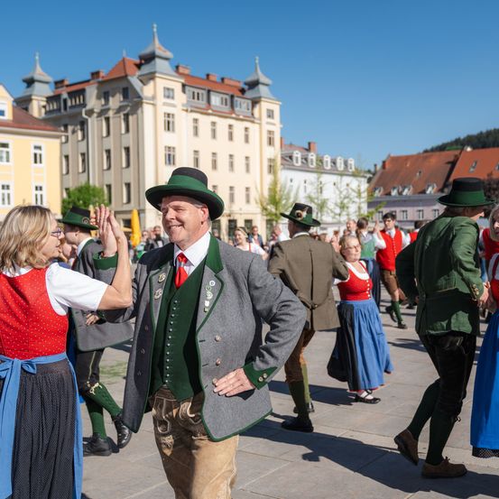 Eine Gruppe von Menschen in traditioneller bayerischer Kleidung tanzt auf der Straße. Gebäude mit mehreren Fenstern und roten Dächern sind im Hintergrund.