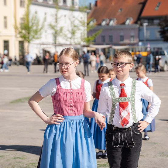 Zwei junge Kinder, ein Mädchen und ein Junge, halten sich an den Händen und gehen auf einer Straße. Sie tragen traditionelle Kleidung. Hinter ihnen laufen mehrere andere Kinder. Gebäude und Bäume sind im Hintergrund.