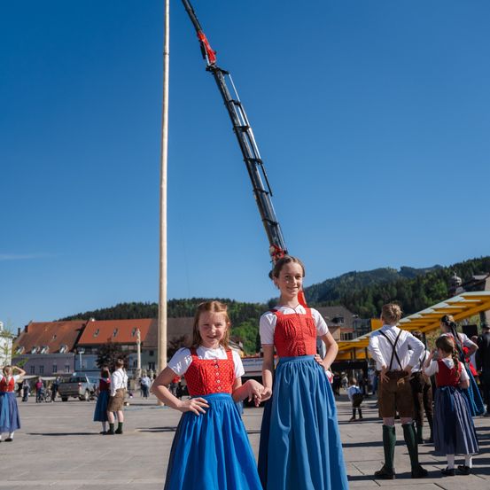 Zwei Mädchen in traditioneller deutscher Kleidung stehen auf einem Stadtplatz, mit einem Kran und Bergen im Hintergrund. Andere Menschen sind ebenfalls in traditioneller Kleidung gekleidet.