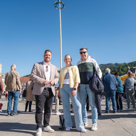 Drei Personen stehen vor einem Pfahl an einem sonnigen Tag. Sie lächeln und schauen in die Kamera. Hinter ihnen stehen mehrere Personen und scheinen zu sprechen. In der Ferne sind Gebäude und Berge zu sehen.