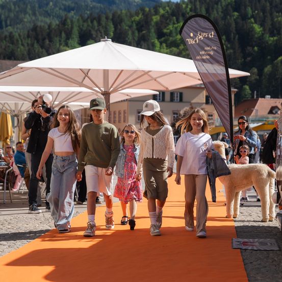 Eine Gruppe von Kindern geht auf einem orangefarbenen Teppich draußen, während ein Fotograf den Moment einfängt. Ein weißer Schirm und ein Banner sind in der Nähe. Der Hintergrund zeigt eine Bergkette.