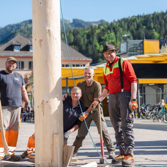 Vier Männer in Arbeitskleidung, einschließlich Handschuhen, sind im Freien. Einer sitzt und hält einen Hammer, während die anderen stehen und lächeln. Ein Holzpfosten steht hinter ihnen, mit Bergen im Hintergrund.