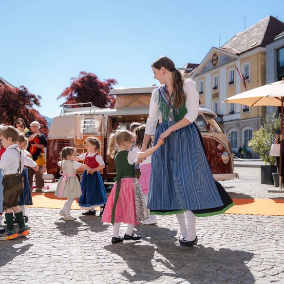 Eine Frau in traditioneller deutscher Kleidung führt eine Gruppe von Kindern auf einem Platz, in der Nähe eines Wohnmobils und eines Gebäudes.