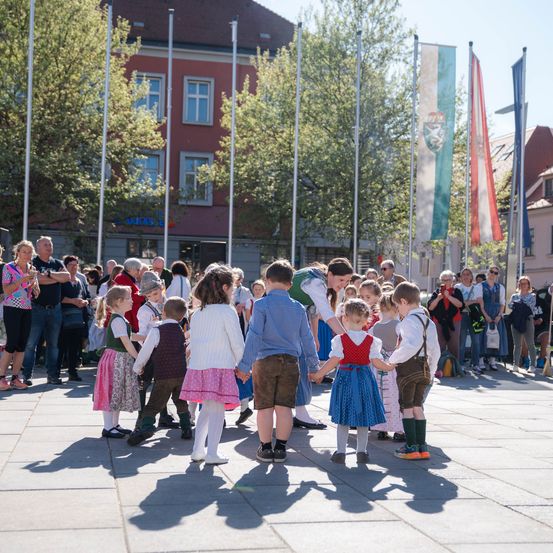 Kinder in traditioneller Kleidung tanzen im Freien in einem Kreis, mit Zuschauern in der Nähe, Gebäude, Bäume und Fahnen im Hintergrund.