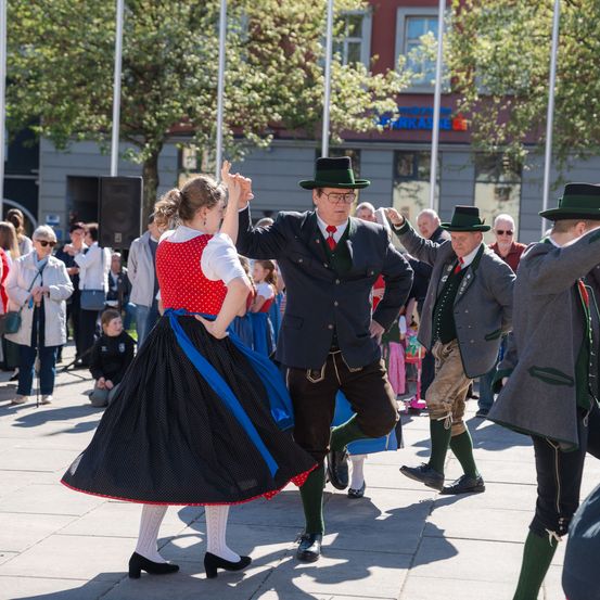 Eine Gruppe von Menschen in traditioneller deutscher Kleidung tanzt auf einem öffentlichen Platz. Sie sind von einer Menge von Zuschauern umgeben.