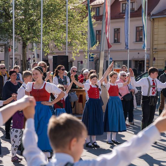 Eine Gruppe von Menschen in traditioneller Kleidung tanzt gemeinsam auf einem Außenbereich, mit Fahnen und Gebäuden im Hintergrund.
