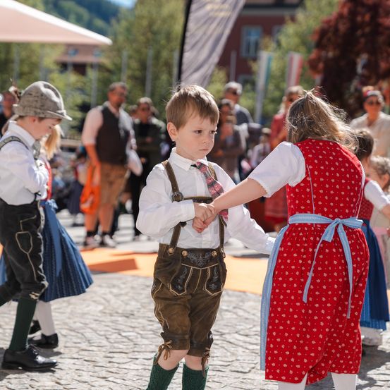 Mehrere Kinder in traditioneller bayerischer Kleidung tanzen auf einer Kopfsteinpflasterstraße. Ein Junge in einem weißen Hemd und braunen Shorts hält die Hände einer Mädchen in einem roten Kleid. Dahinter beobachten weitere Kinder und Erwachsene.