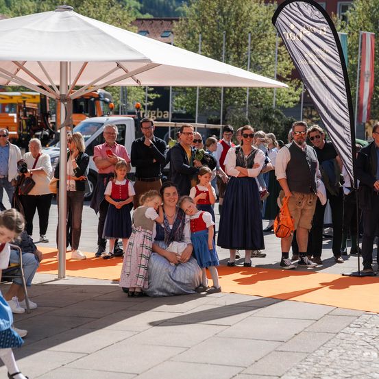 Eine Gruppe von Menschen, einschließlich Kinder und Erwachsene, stehen in einem Kreis auf einem Stadtplatz. Eine Frau in traditioneller Kleidung hält ein Baby, während ein Mann eine Kamera hält. Fahrzeuge und Flaggen sind im Hintergrund.