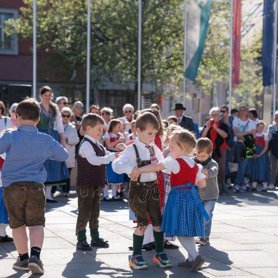 Eine Gruppe von Kindern tanzt auf einem Platz, während Erwachsene zuschauen. Sie tragen traditionelle Kleidung, und im Hintergrund sind Flaggen zu sehen.