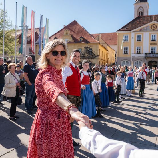 Eine Menschenmenge versammelt sich im Freien, mit Gebäuden im Hintergrund. Eine Frau in einem roten Kleid gibt jemandem in einem weißen Hemd die Hand.