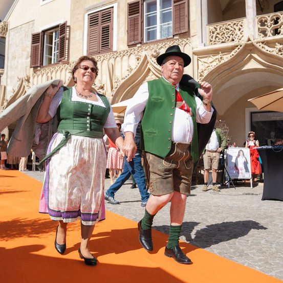 Ein Mann und eine Frau in traditioneller Kleidung gehen auf einem orangefarbenen Teppich vor einem historischen Gebäude. Hinter ihnen gehen Menschen und ein Schirm ist sichtbar.