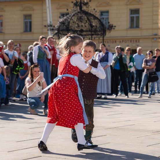 Ein junges Mädchen in einem roten Pünktchenkleid und ein Junge in einem Westchen tanzen gemeinsam auf einem Stadtplatz. Eine Menge beobachtet sie von hinten, einige machen Fotos.