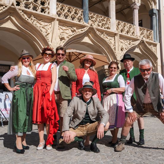 Eine Gruppe von Menschen in traditioneller deutscher Kleidung posiert vor einem historischen Gebäude. Sie lächeln und einige sind gekrümmt oder kniend. Der Hintergrund zeigt einen verzierten Bogen.