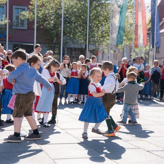 Kinder in traditioneller bayerischer Kleidung tanzen gemeinsam auf einem Stadtplatz während eines Festivals. Zuschauer beobachten von hinten und einige machen Fotos.
