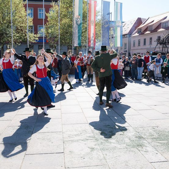 Auf einem Stadtplatz führen Menschen in traditionellen Kostümen einen Tanz im Sonnenlicht auf. Eine Menge sieht von den Seiten zu, mit Fahnen und Gebäuden im Hintergrund.