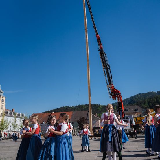 Eine Gruppe von Mädchen in traditioneller Kleidung tanzt um einen Pfahl auf einem Stadtplatz mit Gebäuden, Bäumen und Bergen im Hintergrund.