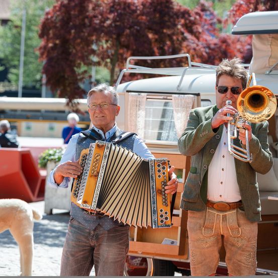Zwei Männer in traditioneller bayerischer Kleidung stehen vor einem Van und spielen Akkordeon und Posaune. Begleitet werden sie von einem Hund und mehreren Zuschauern.