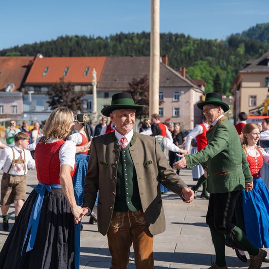 Eine Gruppe von Menschen in traditionellen Kostümen tanzt auf einer Straße vor Gebäuden mit einem Berg im Hintergrund.