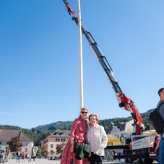 Zwei Frauen stehen neben einem Pfahl mit einem Kran im Hintergrund auf einem Stadtplatz. Ein Lastwagen ist in der Nähe, und es gibt Berge und Gebäude in der Ferne.