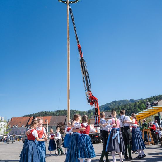 Menschen in traditioneller Kleidung tanzen um einen Pfahl, während im Hintergrund ein großer Kran steht.