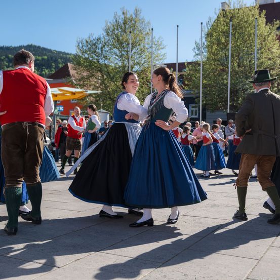 Zwei Frauen tanzen in traditioneller Kleidung auf einem Freigelände, umgeben von Menschen. Ein Mann steht hinter ihnen und schaut zu. In der Ferne gibt es Bäume und Gebäude.