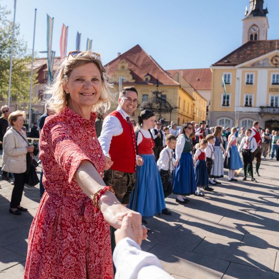Eine Frau in einem roten Kleid lächelt, während sie einem Kind in traditioneller Kleidung die Hand schüttelt, vor einer Menschenmenge im Freien.