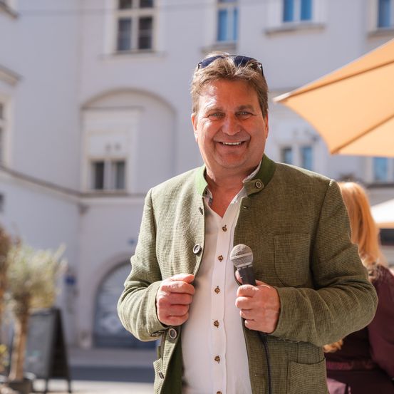 Ein Mann mit einem Mikrofon und einer Jacke lächelt draußen, möglicherweise während einer Rede, mit einem weißen Gebäude und einem Regenschirm im Hintergrund.