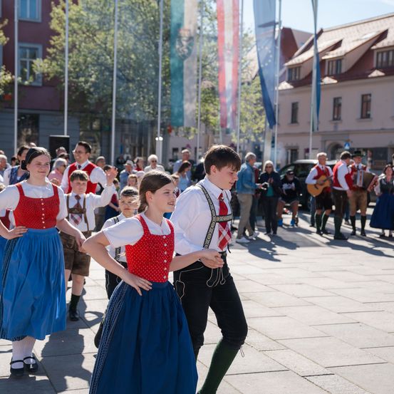 Eine Gruppe von Menschen in traditioneller bayerischer Kleidung tanzt auf einer Kopfsteinpflasterstraße, während andere zusehen und Musiker in der Nähe spielen.