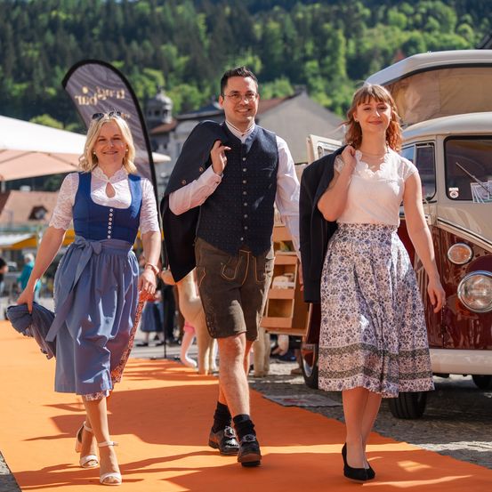 Drei Personen gehen auf einem orangefarbenen Teppich im Freien. Eine Frau in einem blauen Kleid führt, gefolgt von einem Mann in traditioneller Kleidung und einer anderen Frau in einem weißen Kleid. Dahinter steht ein VW-Bulli.