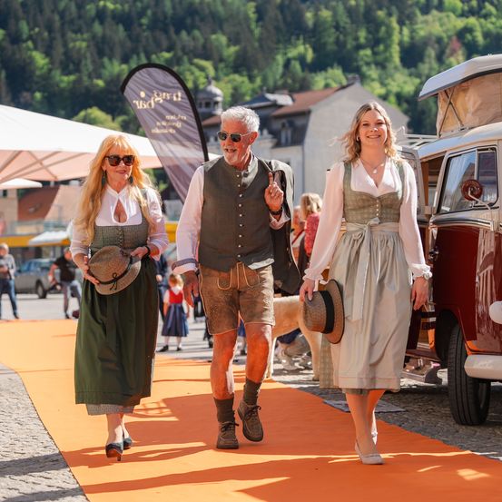Drei Personen in traditioneller bayerischer Tracht gehen auf einem leuchtend orangefarbenen Teppich, mit einem Van und einem Zelt im Hintergrund.