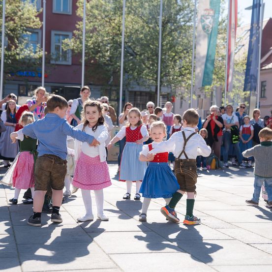 Eine Gruppe von Kindern in traditioneller Kleidung tanzt auf einem öffentlichen Platz, umgeben von einem Publikum.