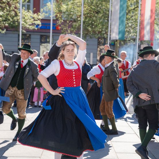 Menschen in traditioneller bayerischer Kleidung tanzen auf einer Stadtstraße. Ein Mann im Vordergrund trägt einen blauen Rock und eine rote Weste. Eine Menge schaut zu.