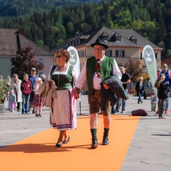 Ein älteres Paar in traditioneller bayerischer Kleidung geht auf einem orangefarbenen Teppich in einer Stadtmitte, mit Häusern und Bergen im Hintergrund.