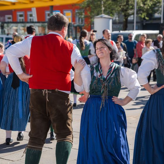 Ein Mann und eine Frau in traditioneller Kleidung tanzen auf einer Straße, während eine Menge Menschen zusieht.