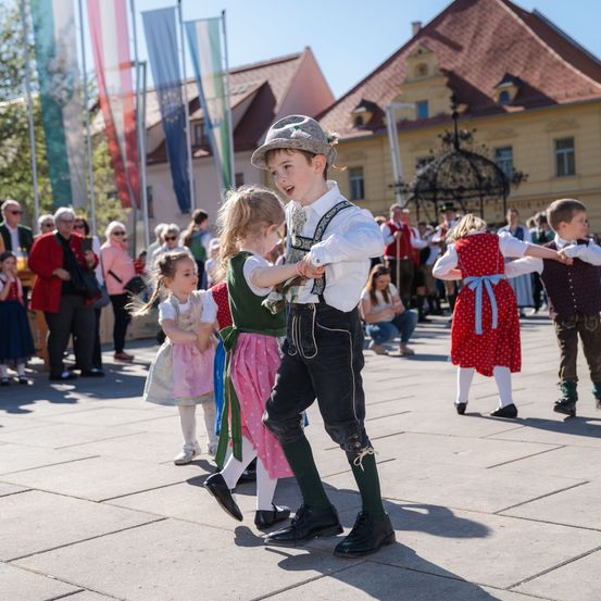 Kinder in traditioneller Kleidung tanzen auf einem Stadtplatz, während Erwachsene im Hintergrund stehen und Fahnen wehen.