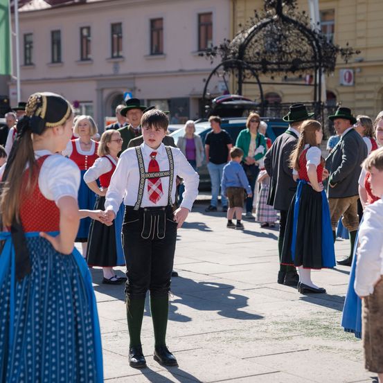 Eine Gruppe von Menschen, einschließlich Kindern, gekleidet in traditioneller bayerischer Tracht, steht auf einem Stadtplatz. Ein Junge hält die Hand einer Mädchen. Hinter ihnen gehen Erwachsene umher, einige tragen Hüte. Im Hintergrund sind Gebäude und Autos zu sehen.