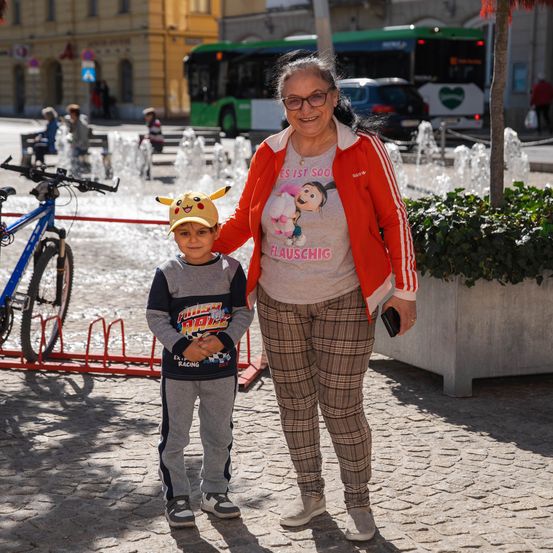 Eine Frau und ein kleiner Junge stehen lächelnd nebeneinander auf einem Stadtplatz. Der Junge trägt einen Pikachu-Hut. Hinter ihnen ist ein Springbrunnen und ein Fahrrad zu sehen.